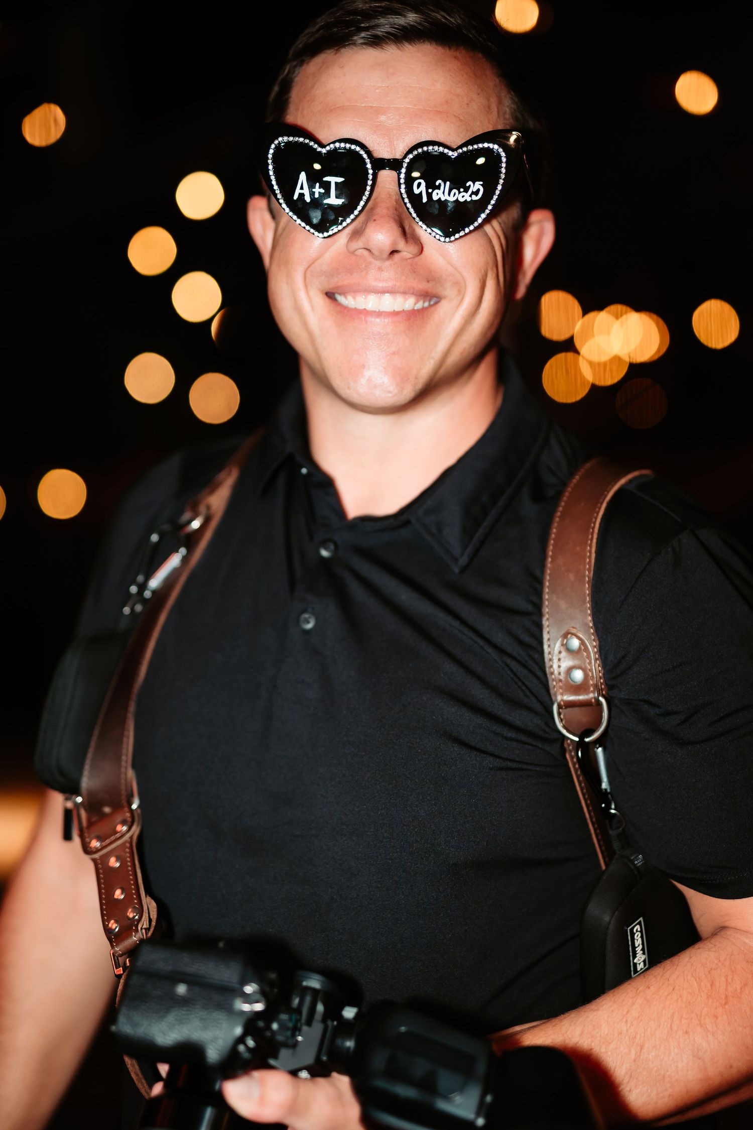 Person wearing black shirt and leather harness smiles in dimly lit setting with bokeh lights in background.