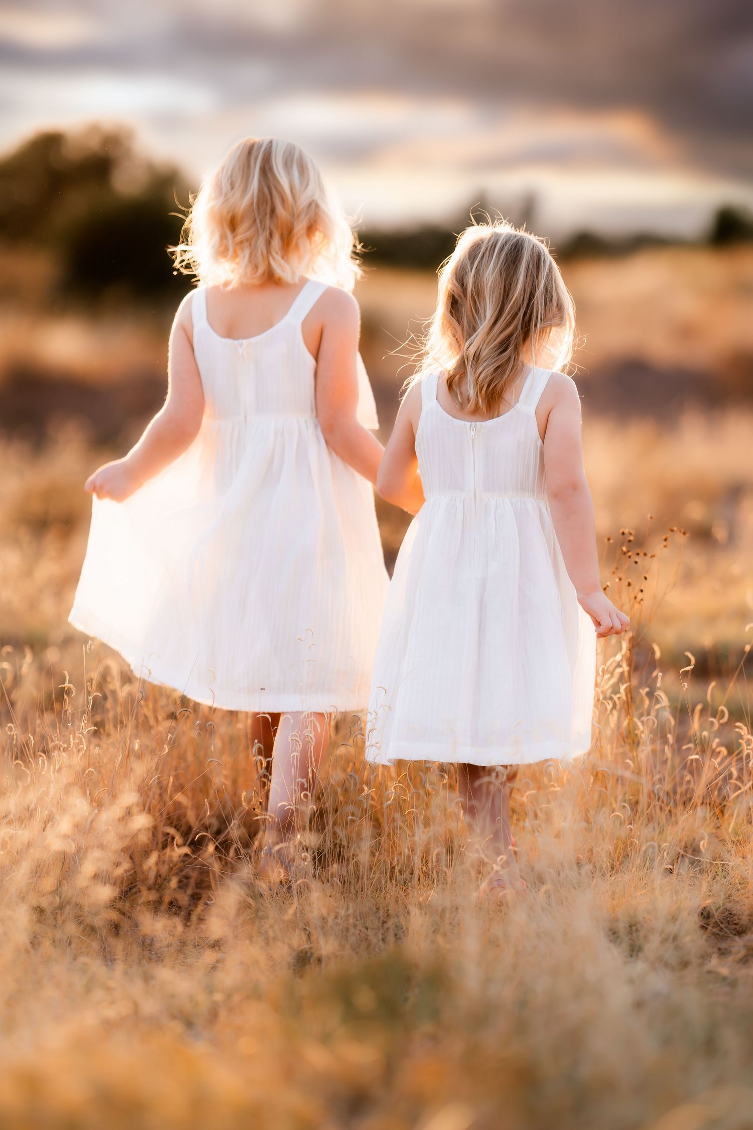 Two young girls in white dresses walk together through a golden field at sunset creating a dreamy summer scene.