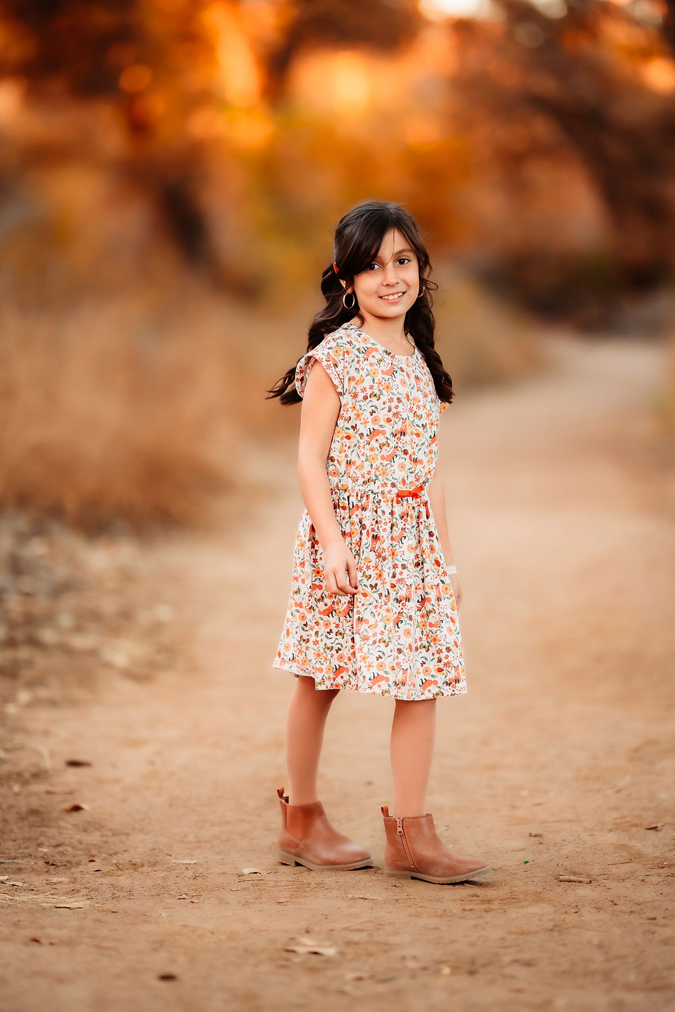 Young girl in floral dress and brown boots smiling on a dirt path with warm autumn bokeh background.