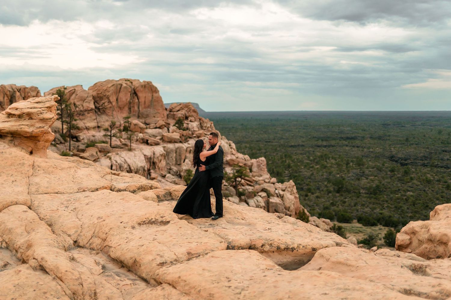 Couple in black attire embracing on red rock formations with vast green landscape and cloudy sky backdrop.