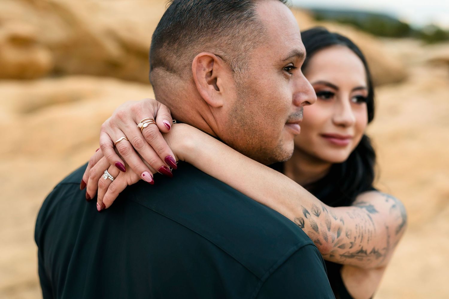 Close-up of couple embracing, woman with ring and tattoo visible wrapping arms around mans neck outdoors.