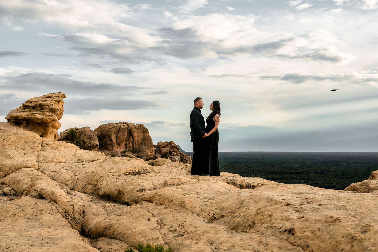 Couple in black attire standing on golden sandstone rocks with vast landscape and dramatic cloudy sky behind them.