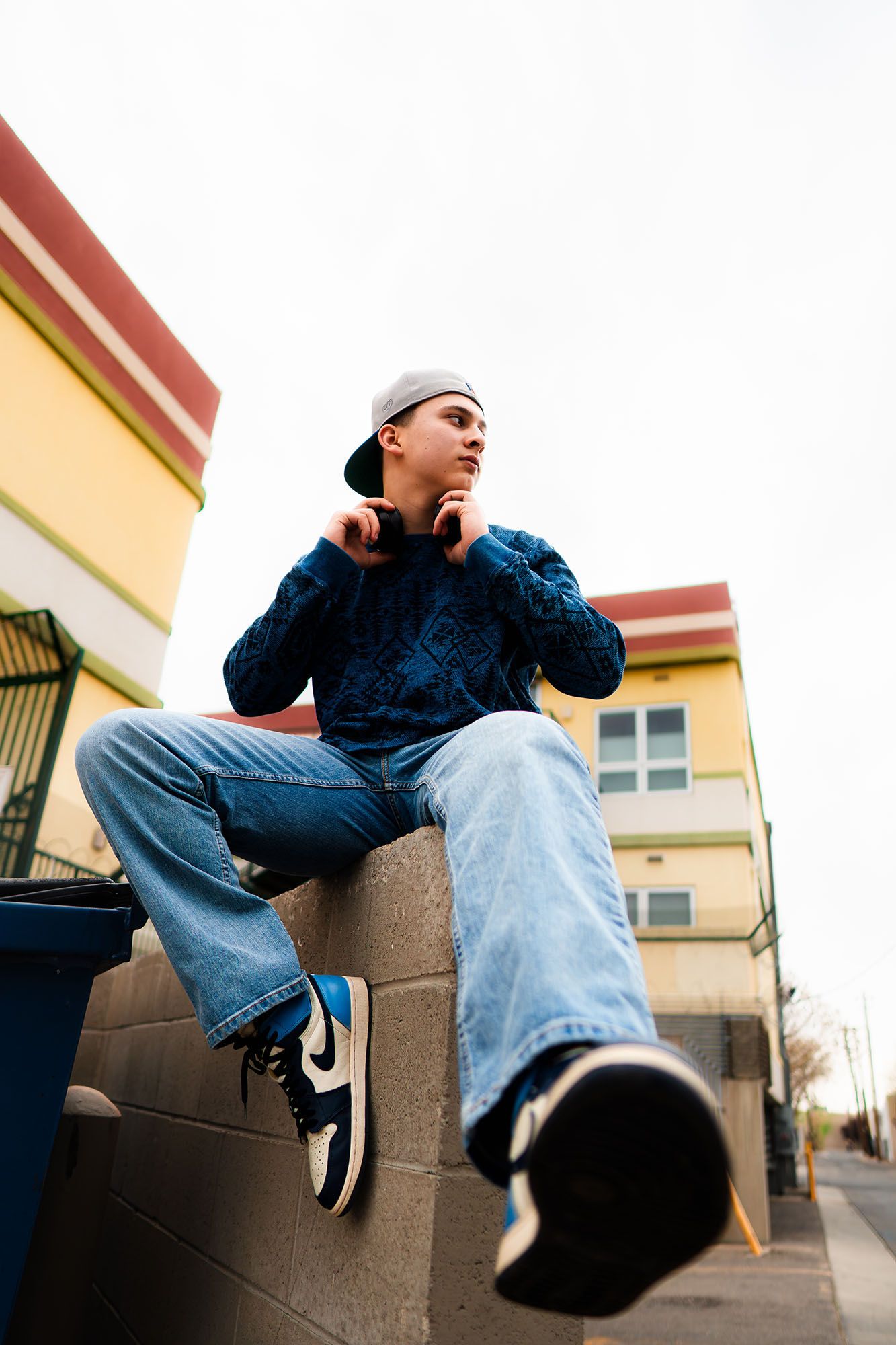 Teen boy in blue sweater, jeans, and Nike Air Jordans sitting on concrete wall outdoors.