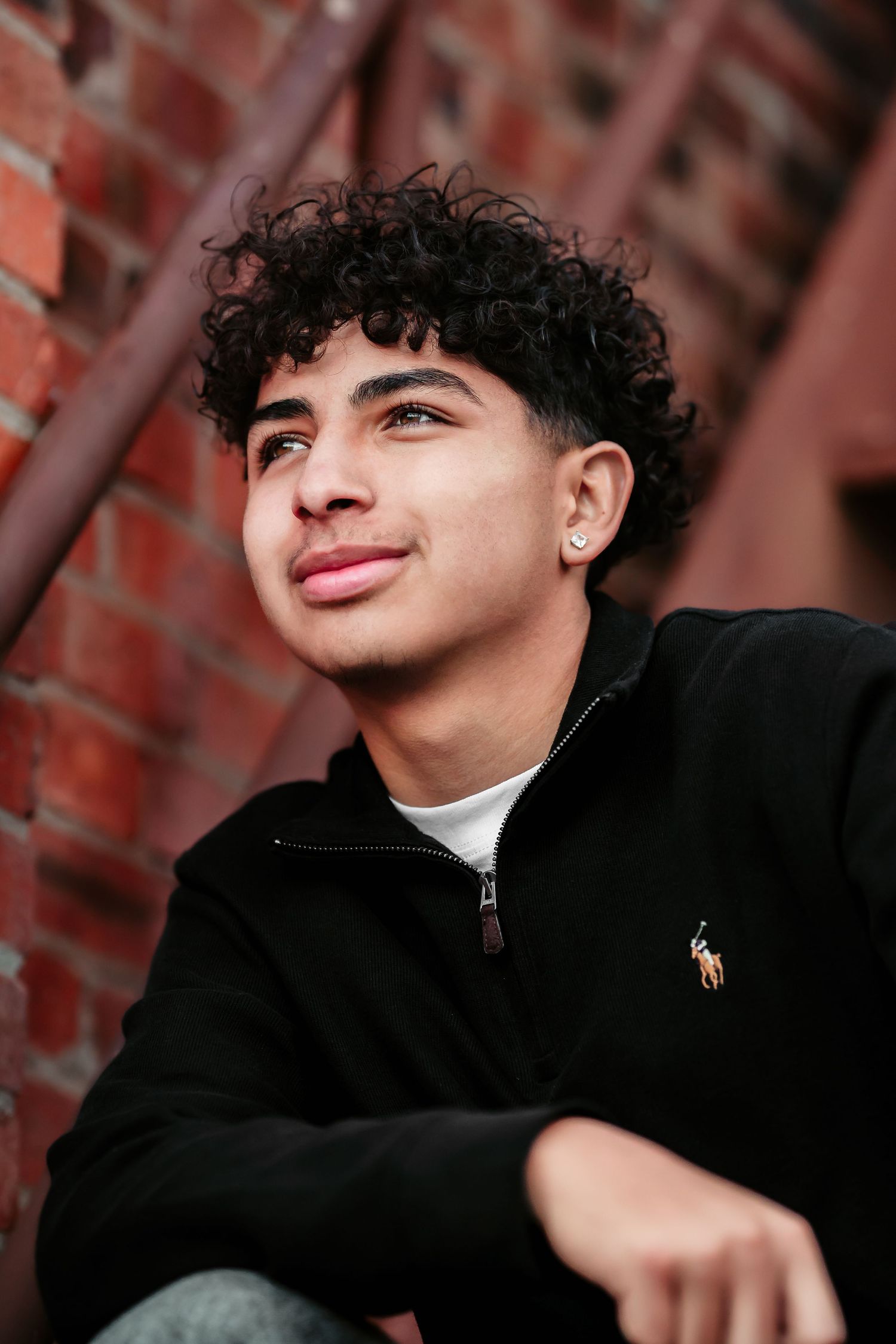 Portrait of a young person with curly black hair and an earring wearing a black Polo zip-up against a brick wall background.