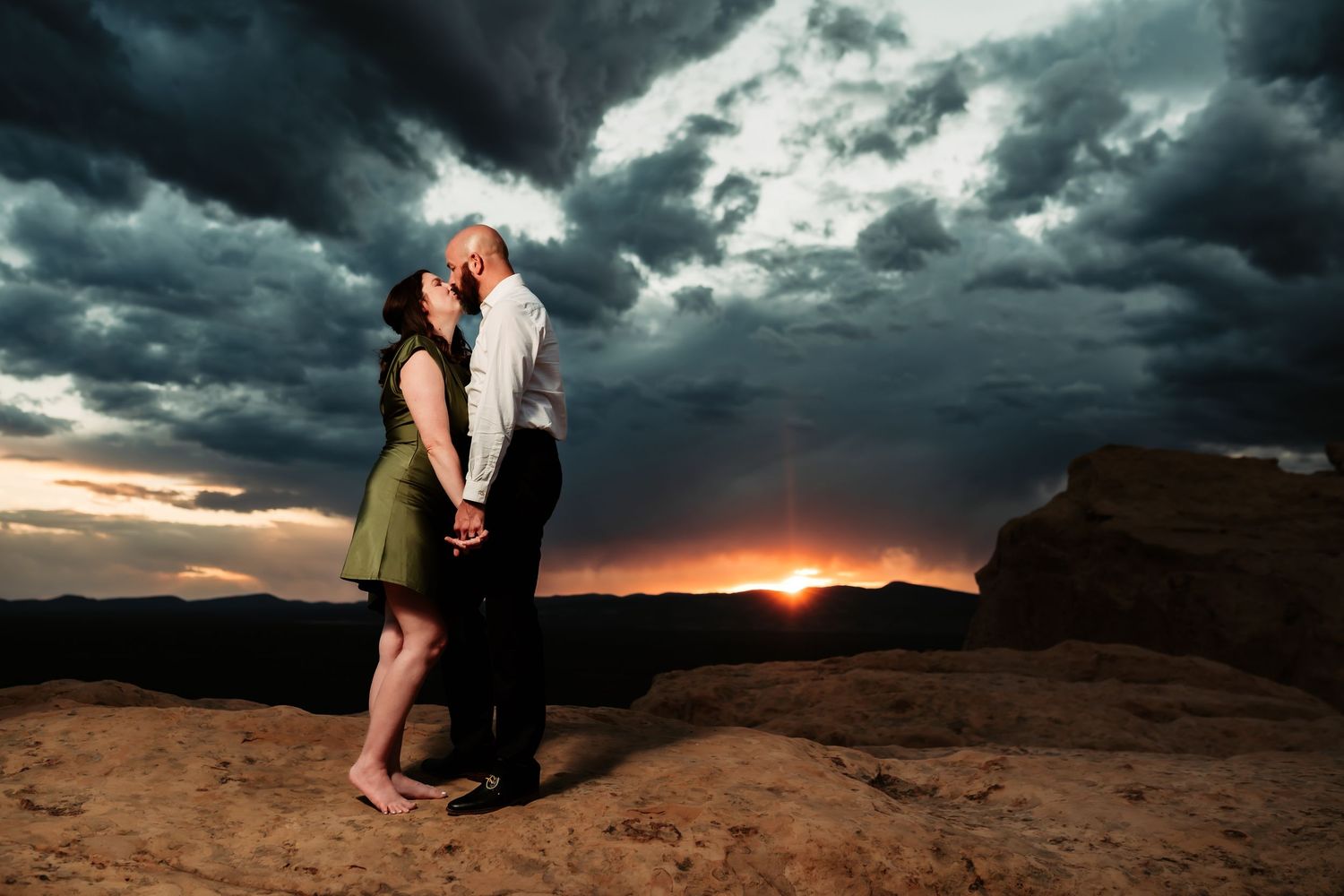 Two people embrace on a desert cliff at sunset beneath dramatic storm clouds in the background.