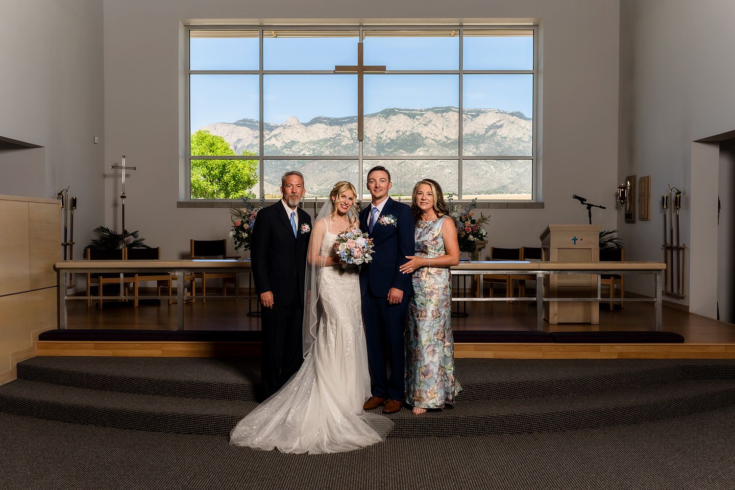 Bride and groom pose with family at church altar, mountain view through window, wedding ceremony venue.