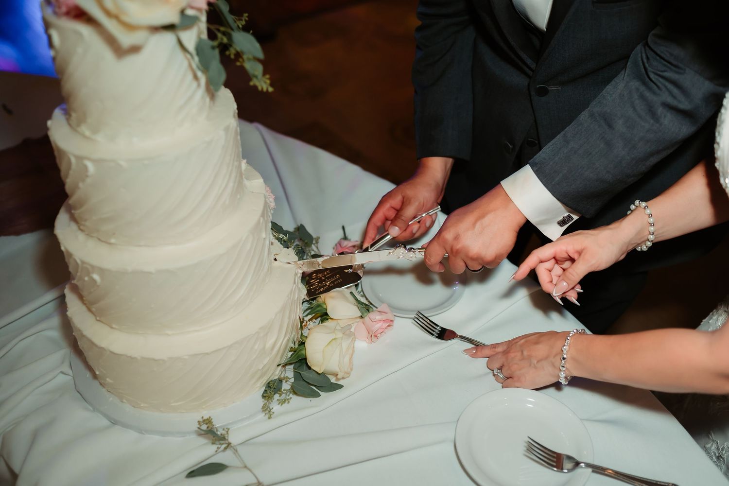 Close-up shot of hands cutting into a white tiered wedding cake decorated with flowers.