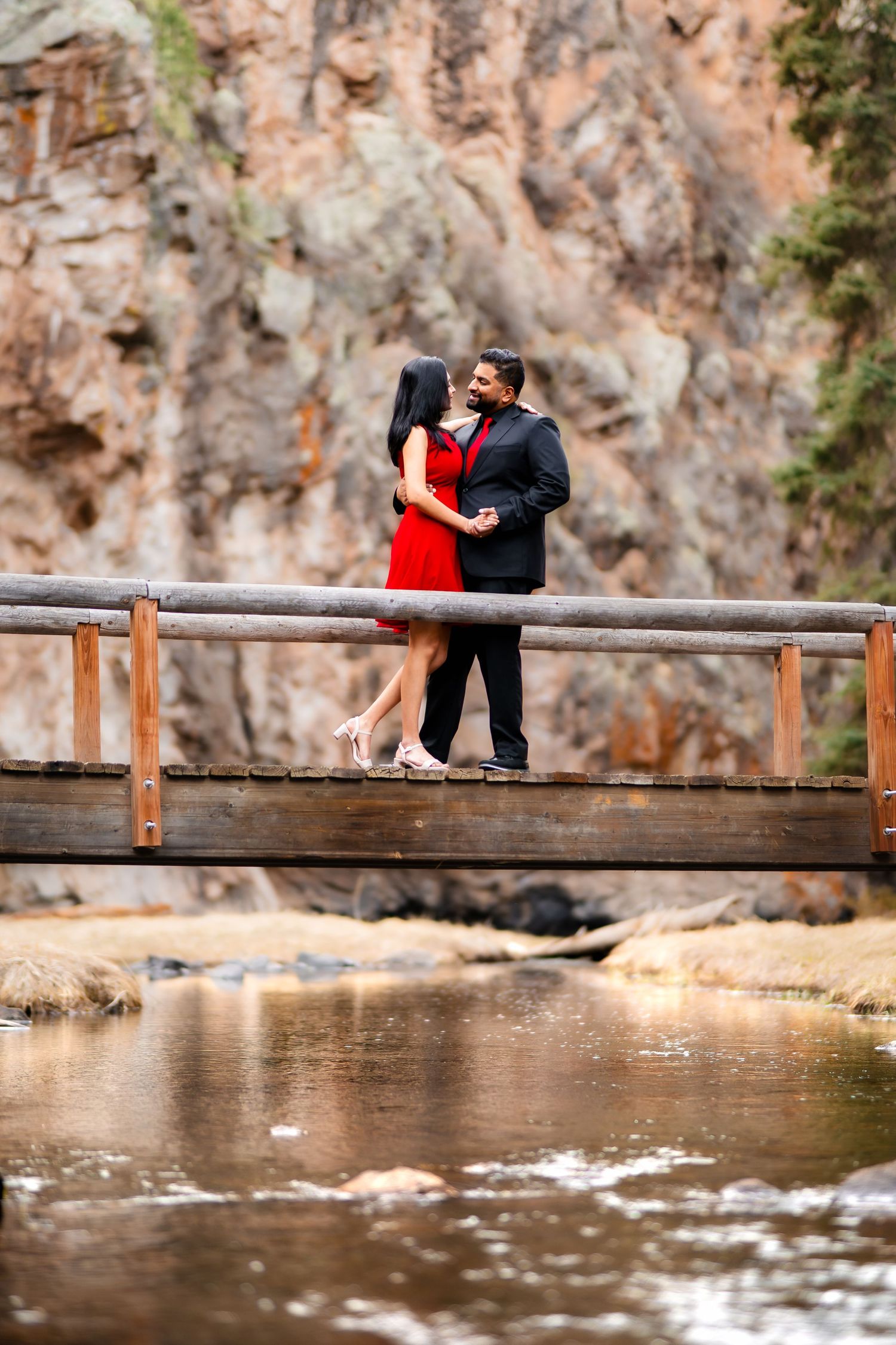 Couple stands together on wooden footbridge over mountain stream with red rock cliffs behind them.