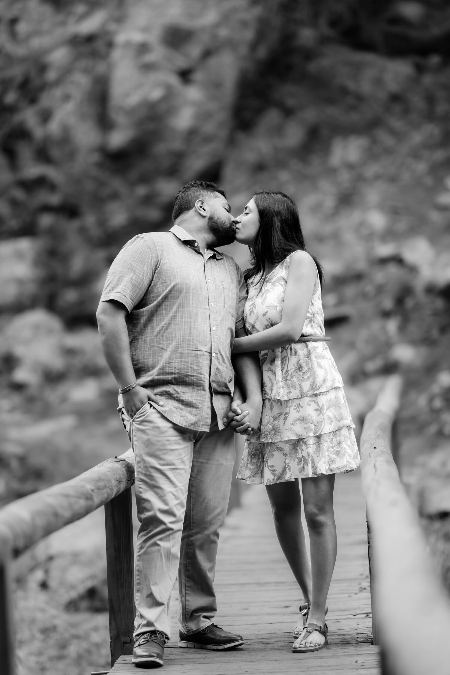 A romantic black and white photograph of a couple sharing a kiss on a wooden footbridge in nature.