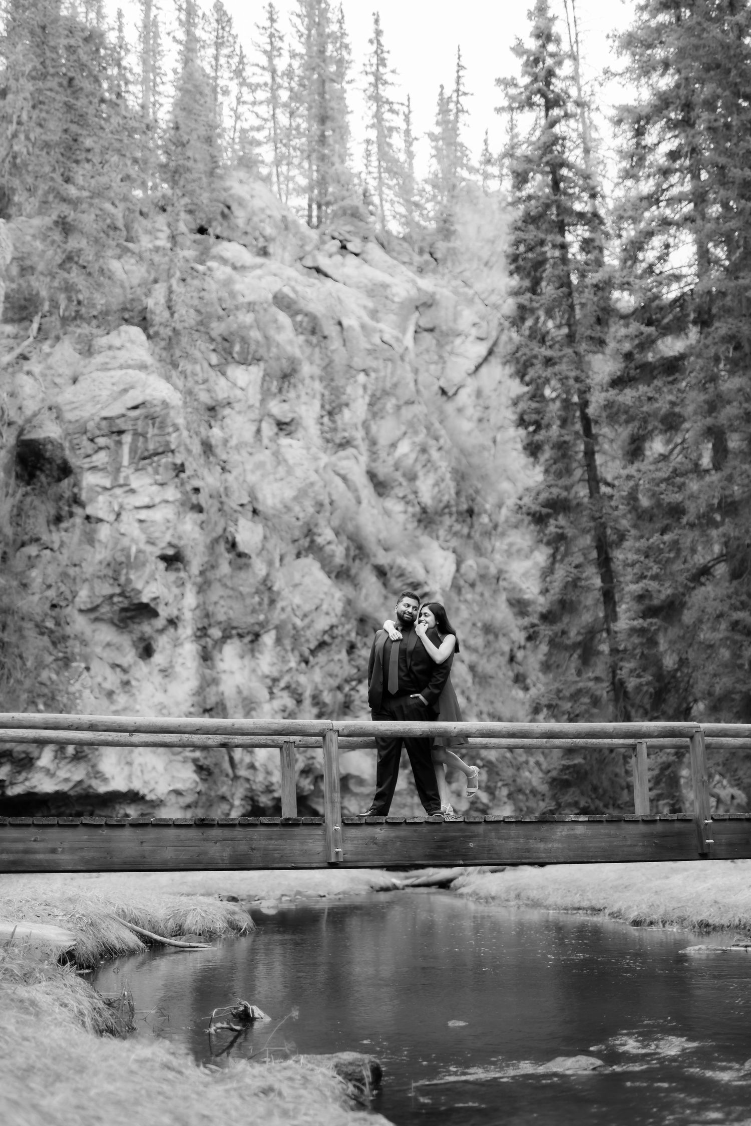 Silhouetted couple embracing on bridge with dramatic mountain landscape and evergreen trees.
