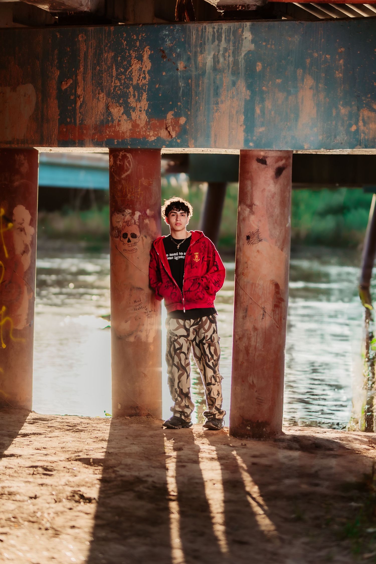 Person in red jacket and camo pants standing between concrete pillars near water.