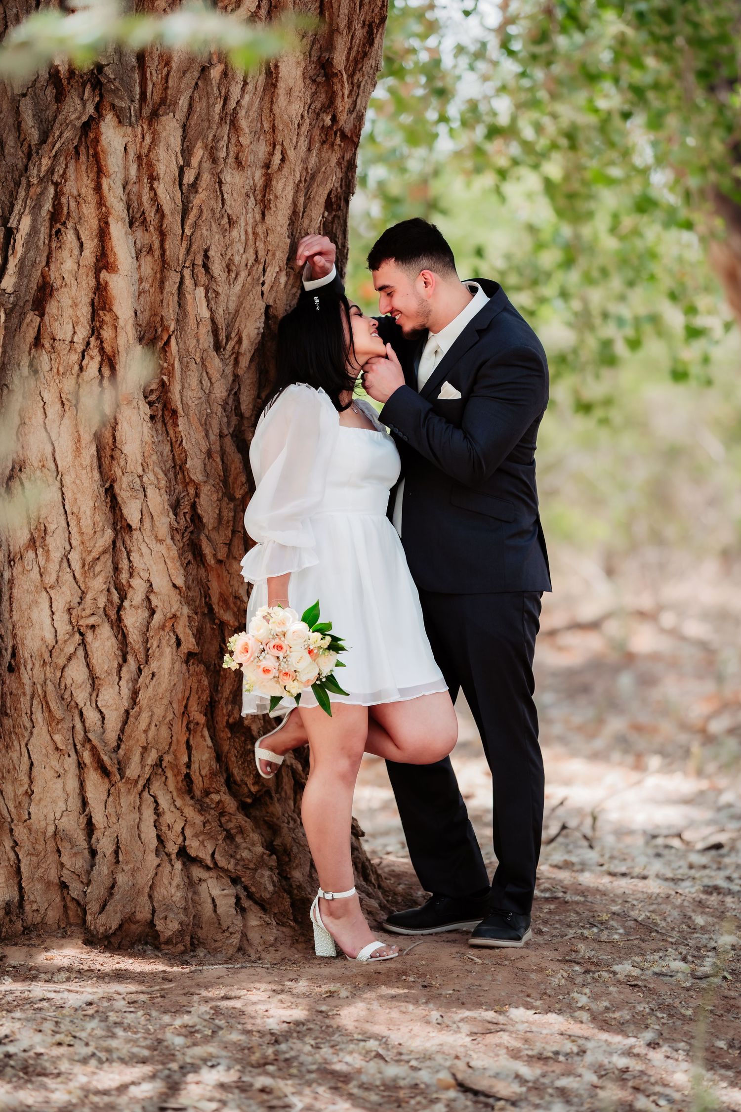 A romantic wedding couple shares an intimate moment against a large tree trunk while holding a pink bouquet.