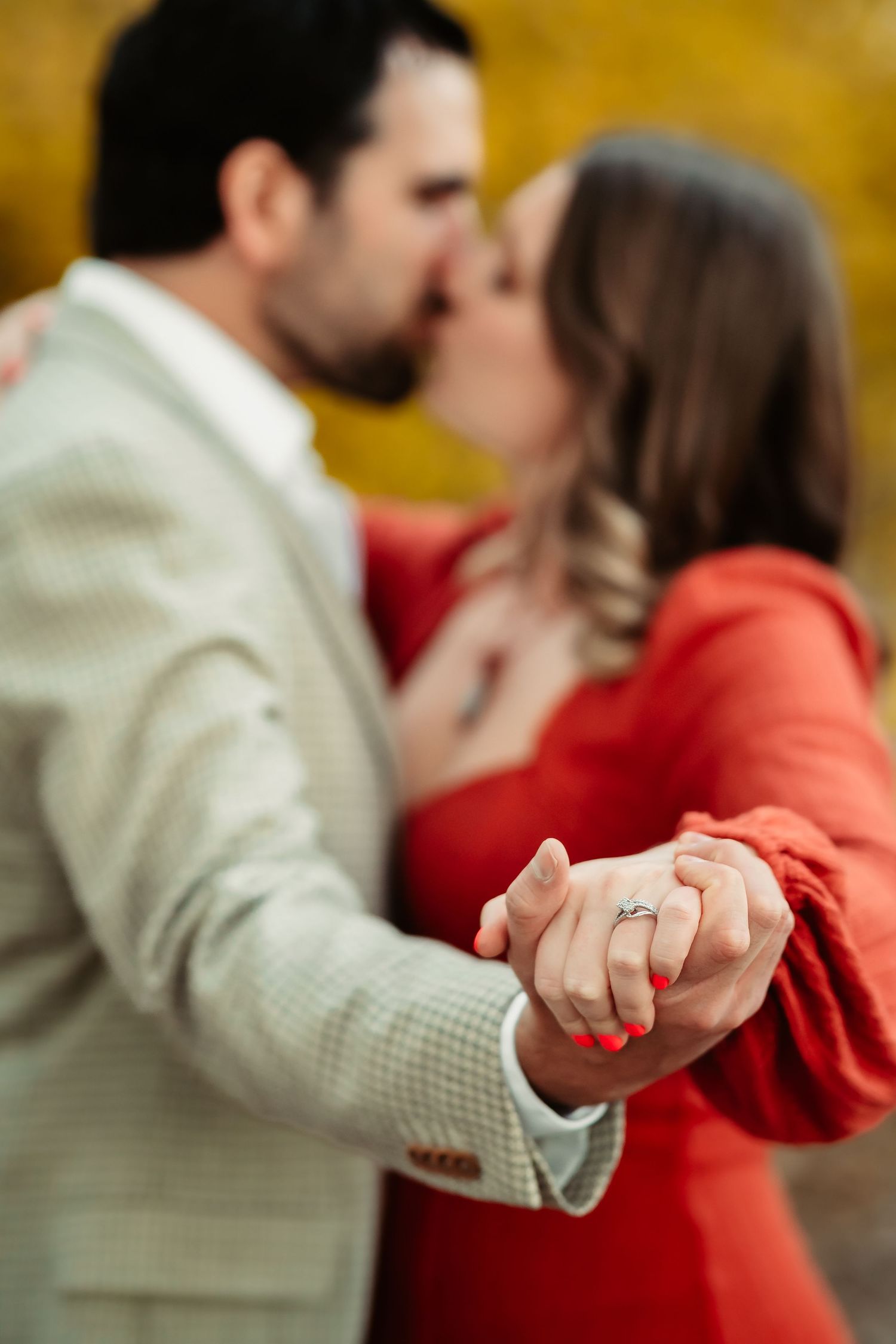 A couple in a white shirt and red sweater hold hands intimately against a golden autumn background.