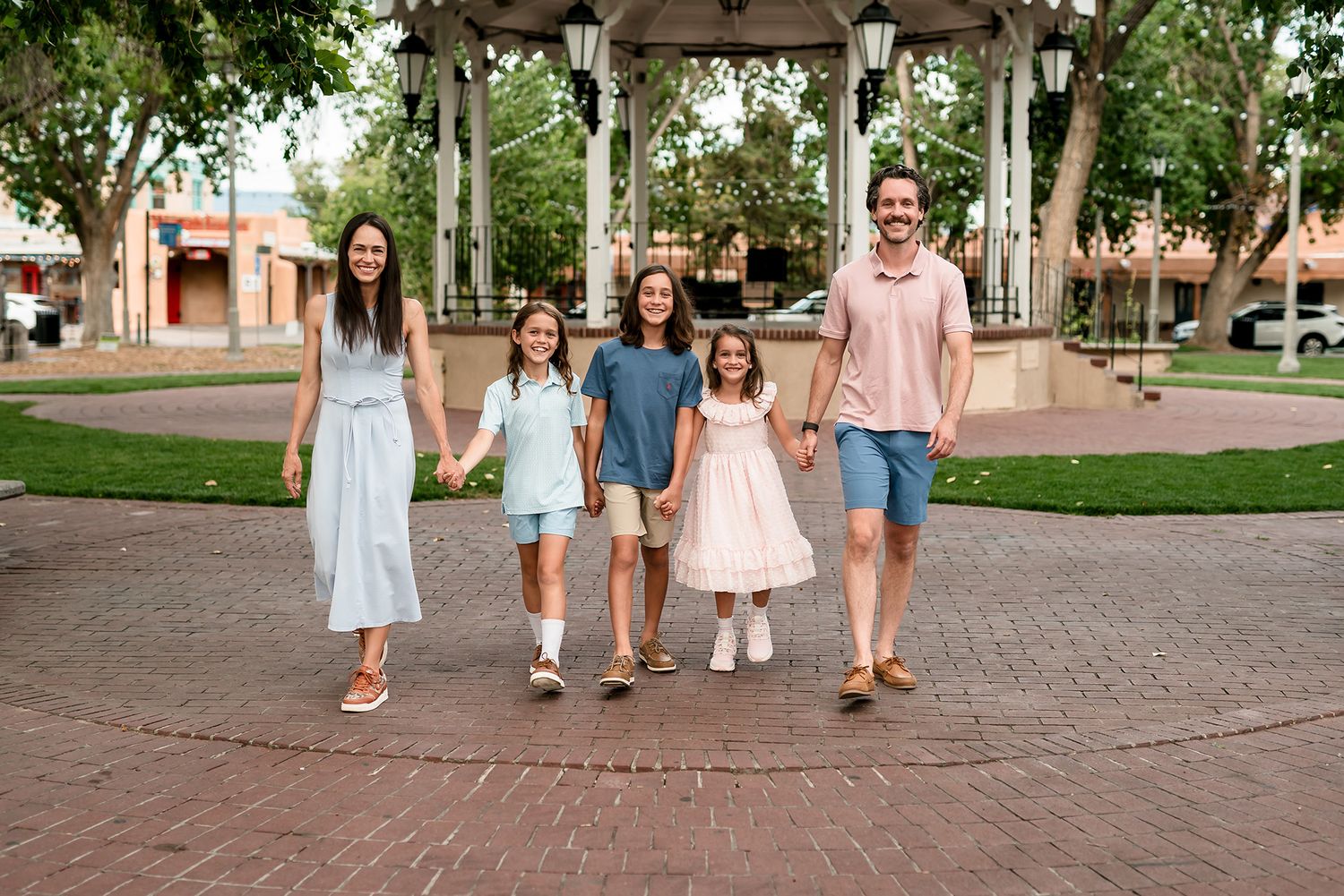 Happy family of five walking together in a brick plaza near a gazebo, dressed in pastel summer outfits.