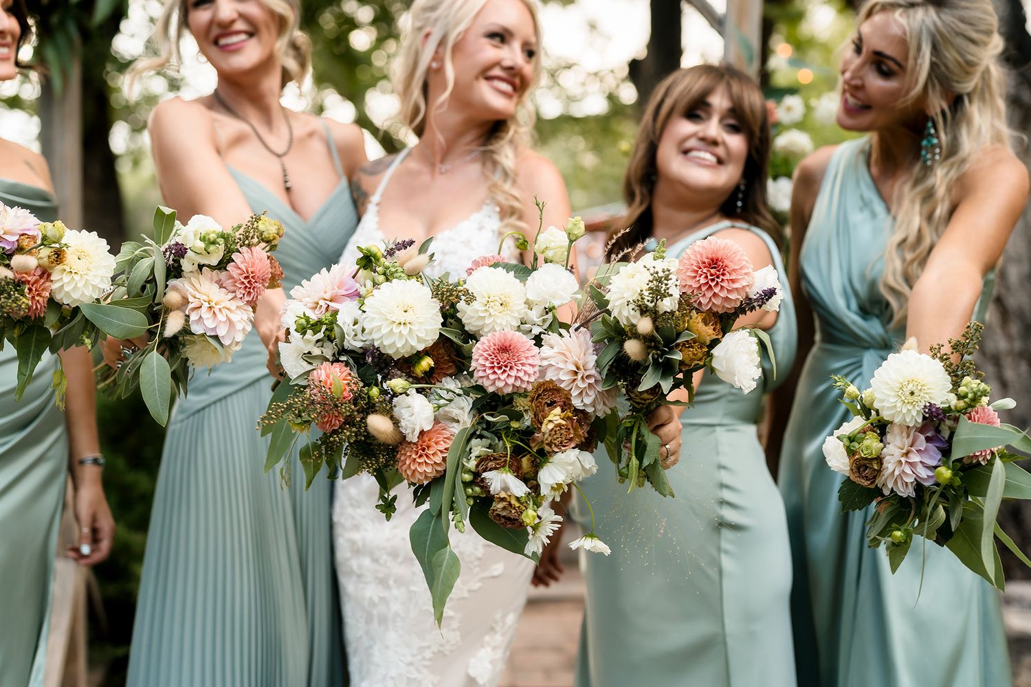 Bride in white lace gown with bridesmaids in sage green dresses holding colorful floral bouquets outdoors.