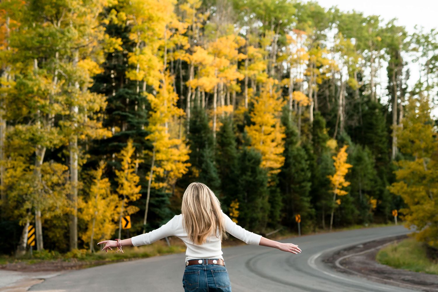 Blonde woman with arms outstretched standing on road surrounded by vibrant yellow autumn aspen trees.