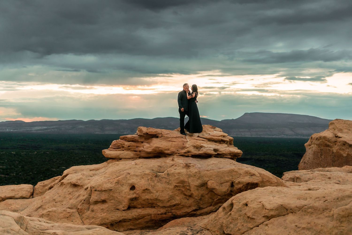 A couple embraces on rugged sandstone cliffs under a moody stormy sky with sweeping desert views.