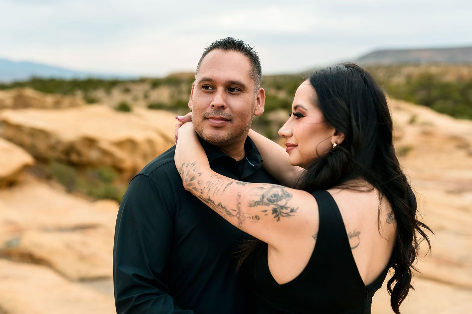 Couple embracing in desert landscape, woman with floral tattoo wrapping arm around man's neck.