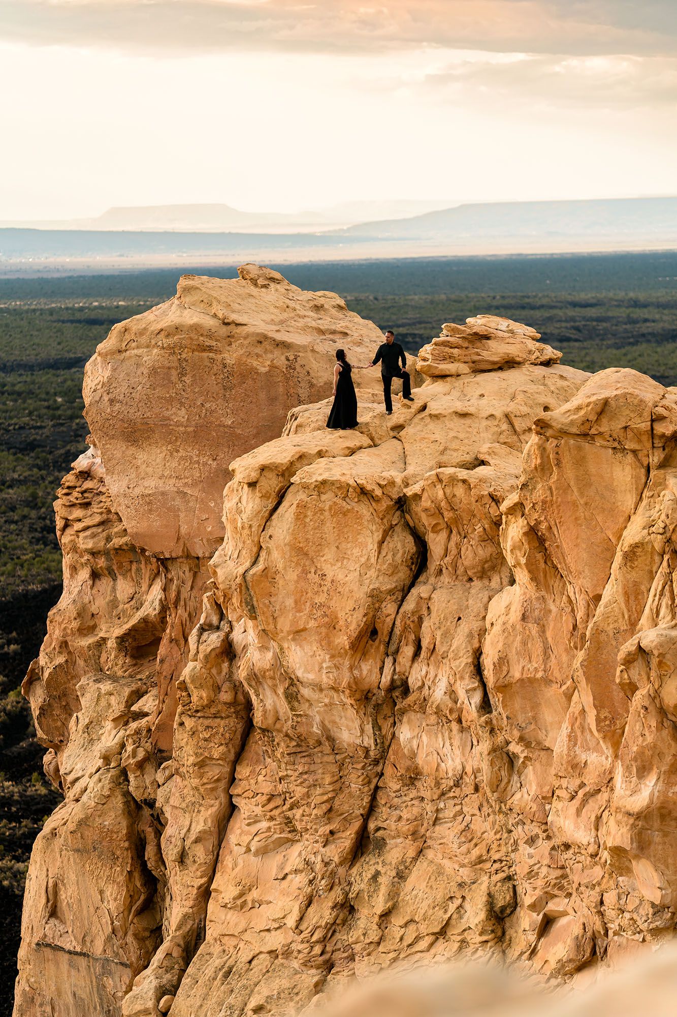 Two figures in black stand atop a dramatic sandstone cliff overlooking a vast desert landscape at sunset.