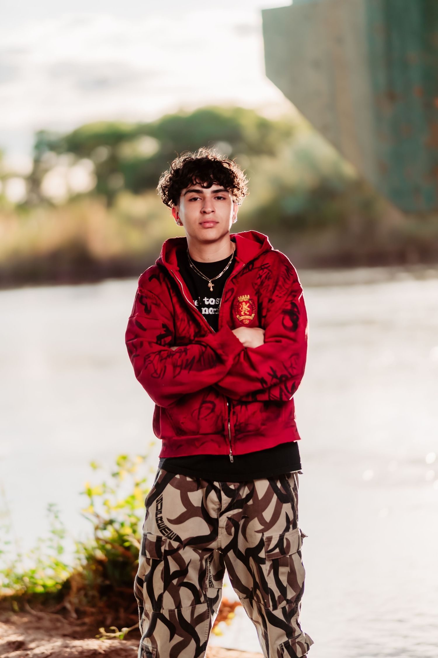 Person in a red hoodie and patterned pants poses outdoors with sunlit background.