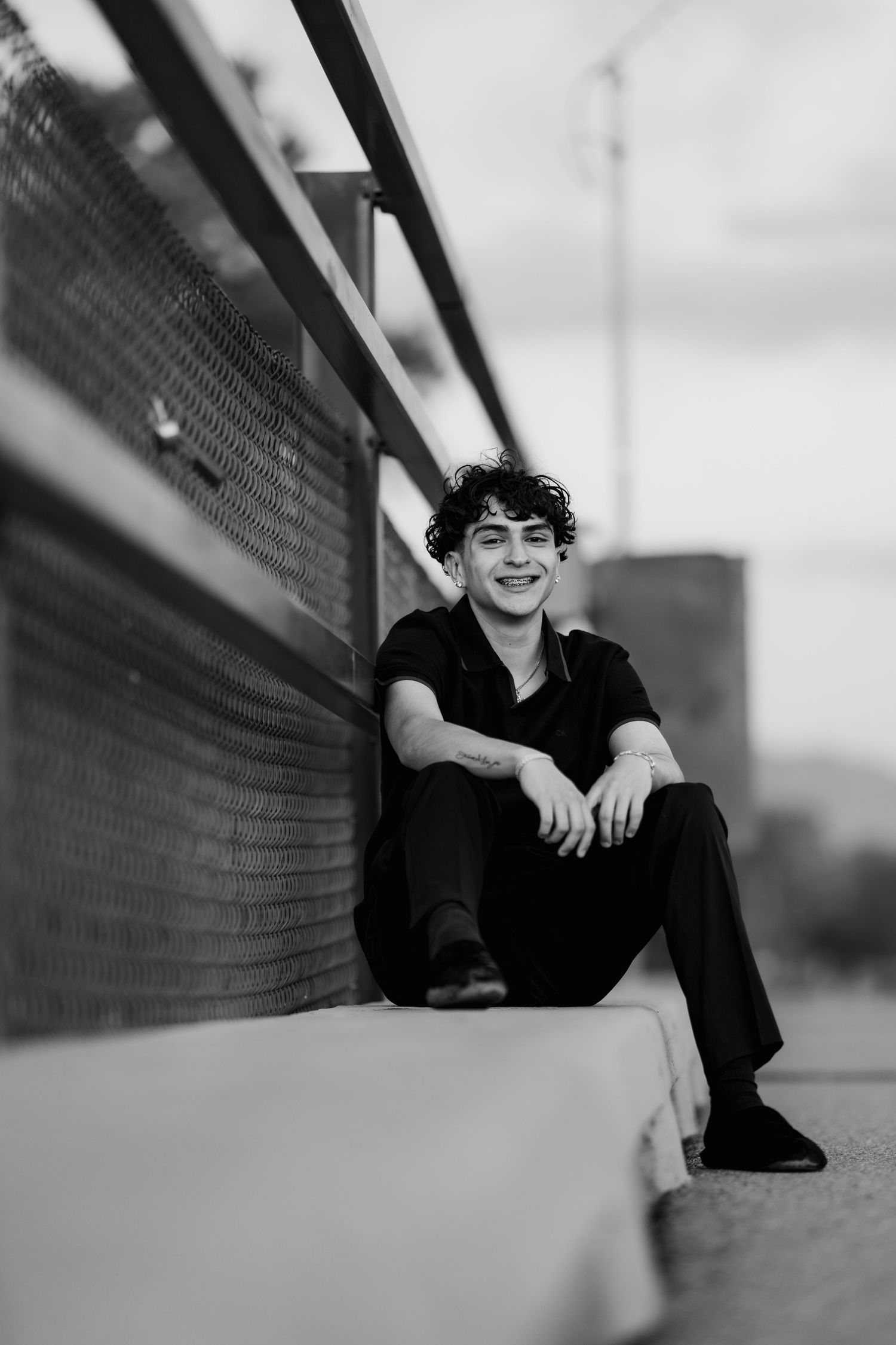 A candid black and white portrait of someone sitting casually against a fence with an urban backdrop.