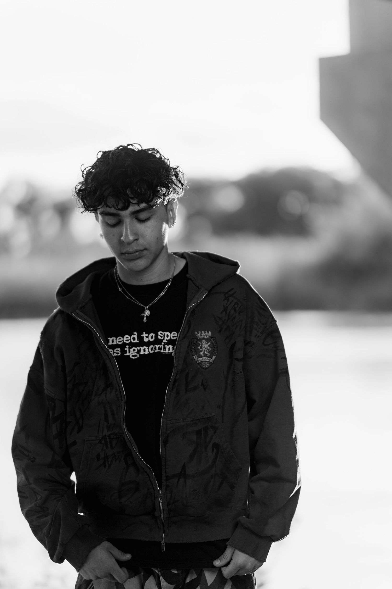 A person in a dark hoodie with curly hair looks down thoughtfully in black and white portrait style.