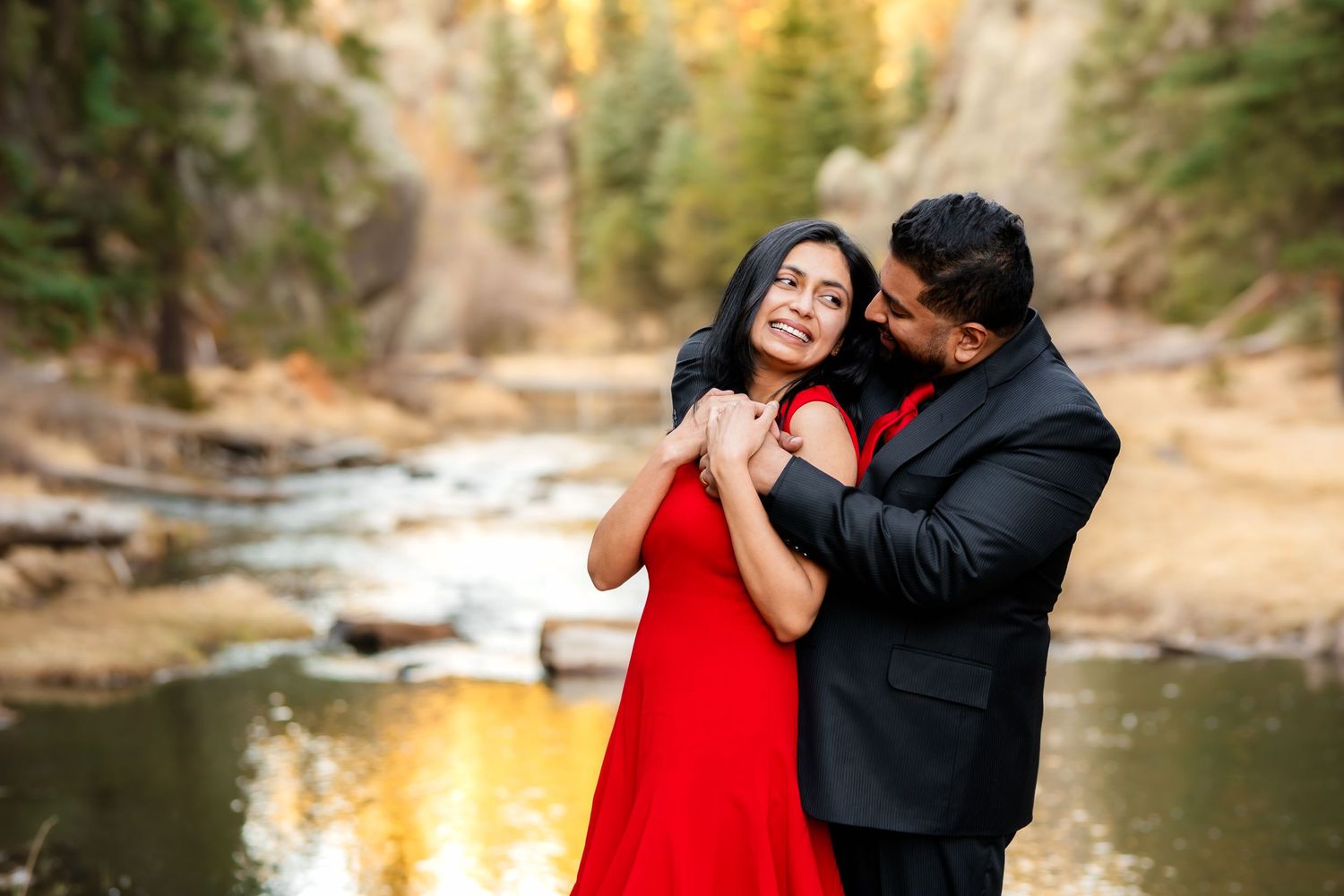 Romantic couple embraces by a serene mountain stream with sunlit cliffs in the background.