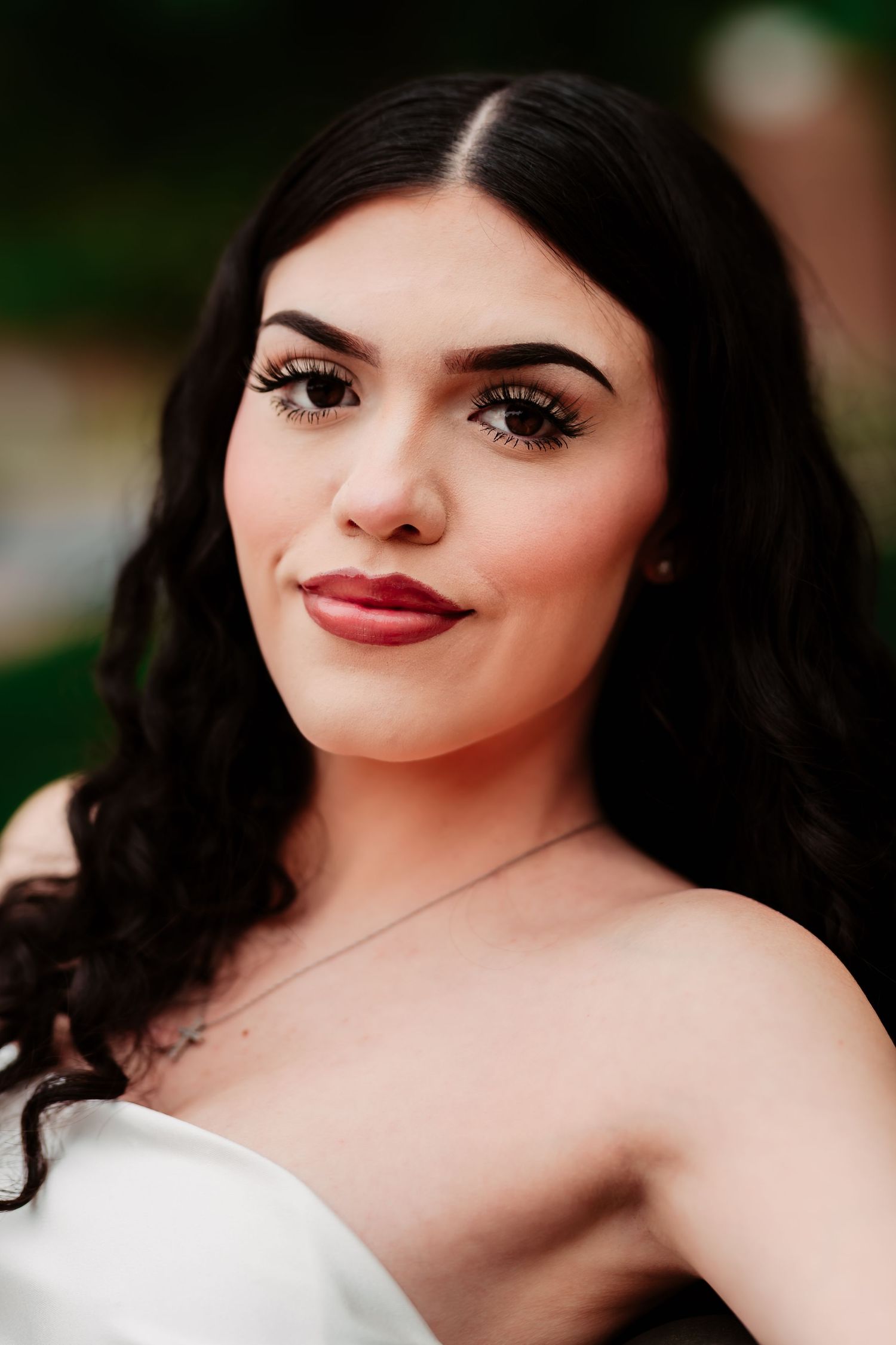 Portrait with dark wavy hair wearing red lipstick and a white strapless top against blurred outdoor background.
