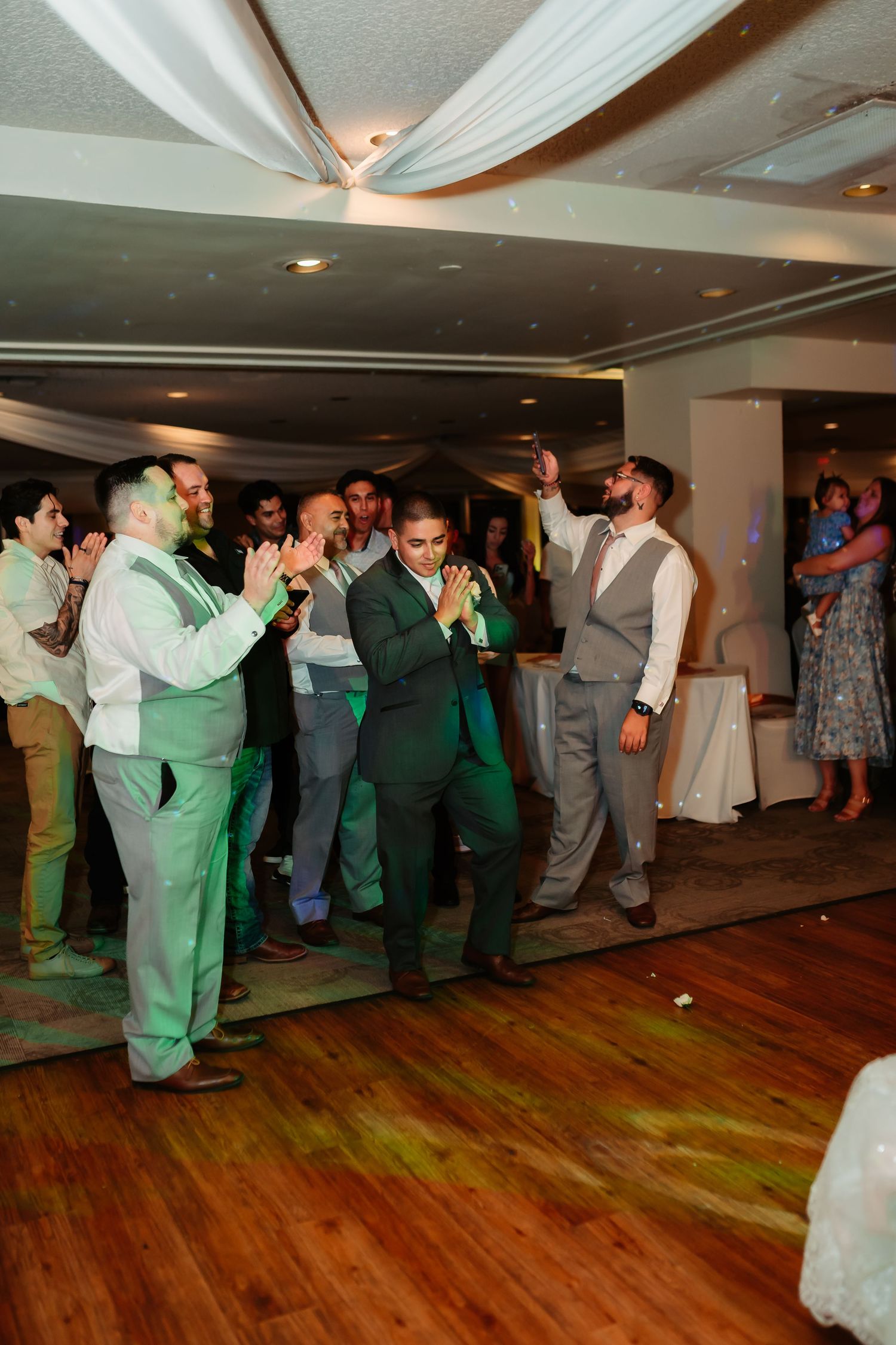 Wedding guests in formal attire dancing and celebrating on a wooden dance floor at a reception venue.