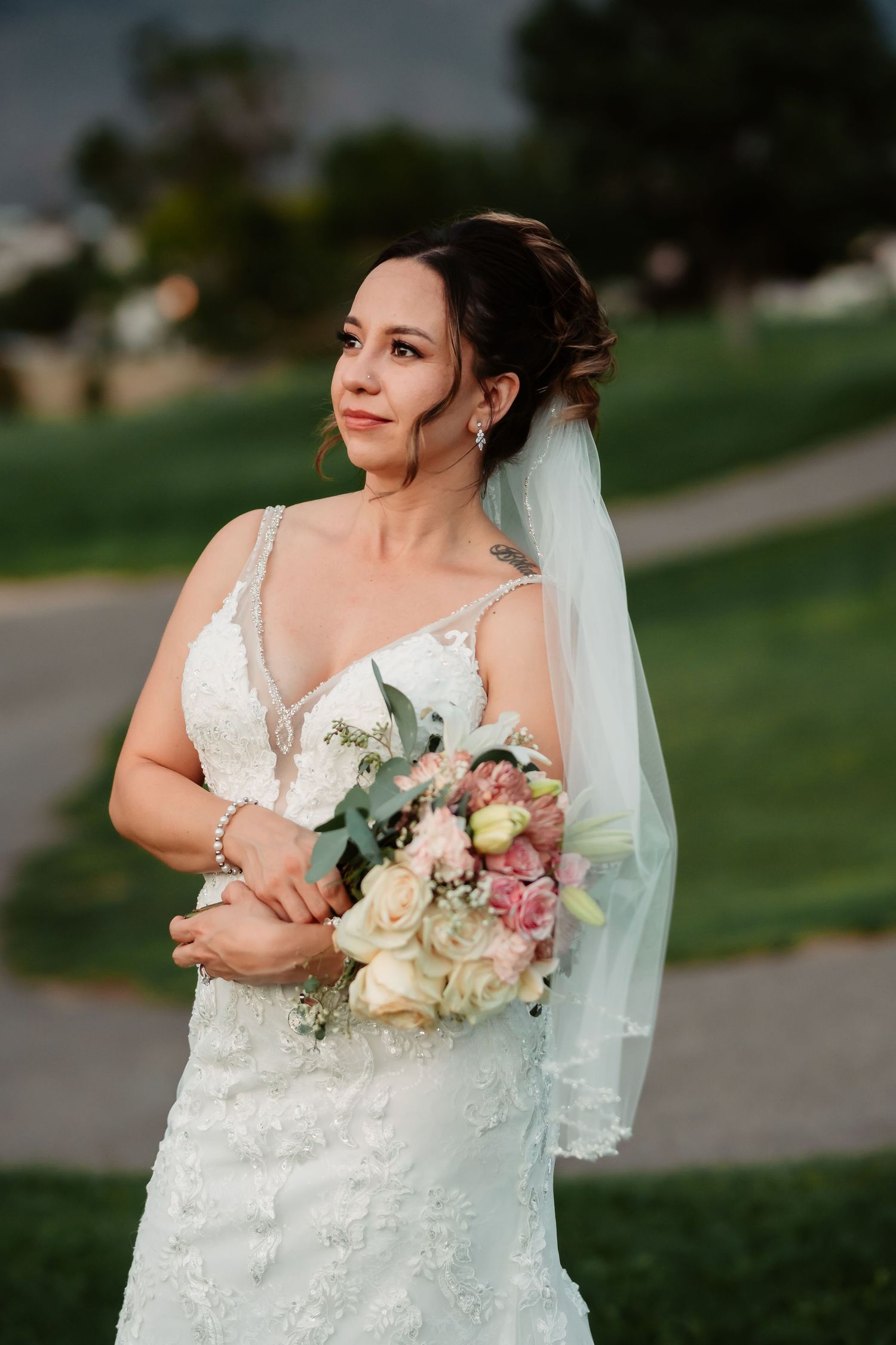 A bride in a lace wedding dress holds a pink and white bouquet against a green outdoor backdrop.