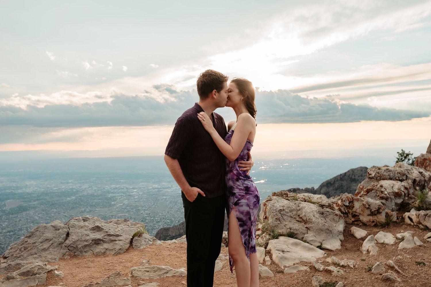 Romantic couple sharing a kiss at sunset on a mountaintop overlook with dramatic cloudy skies in the background.
