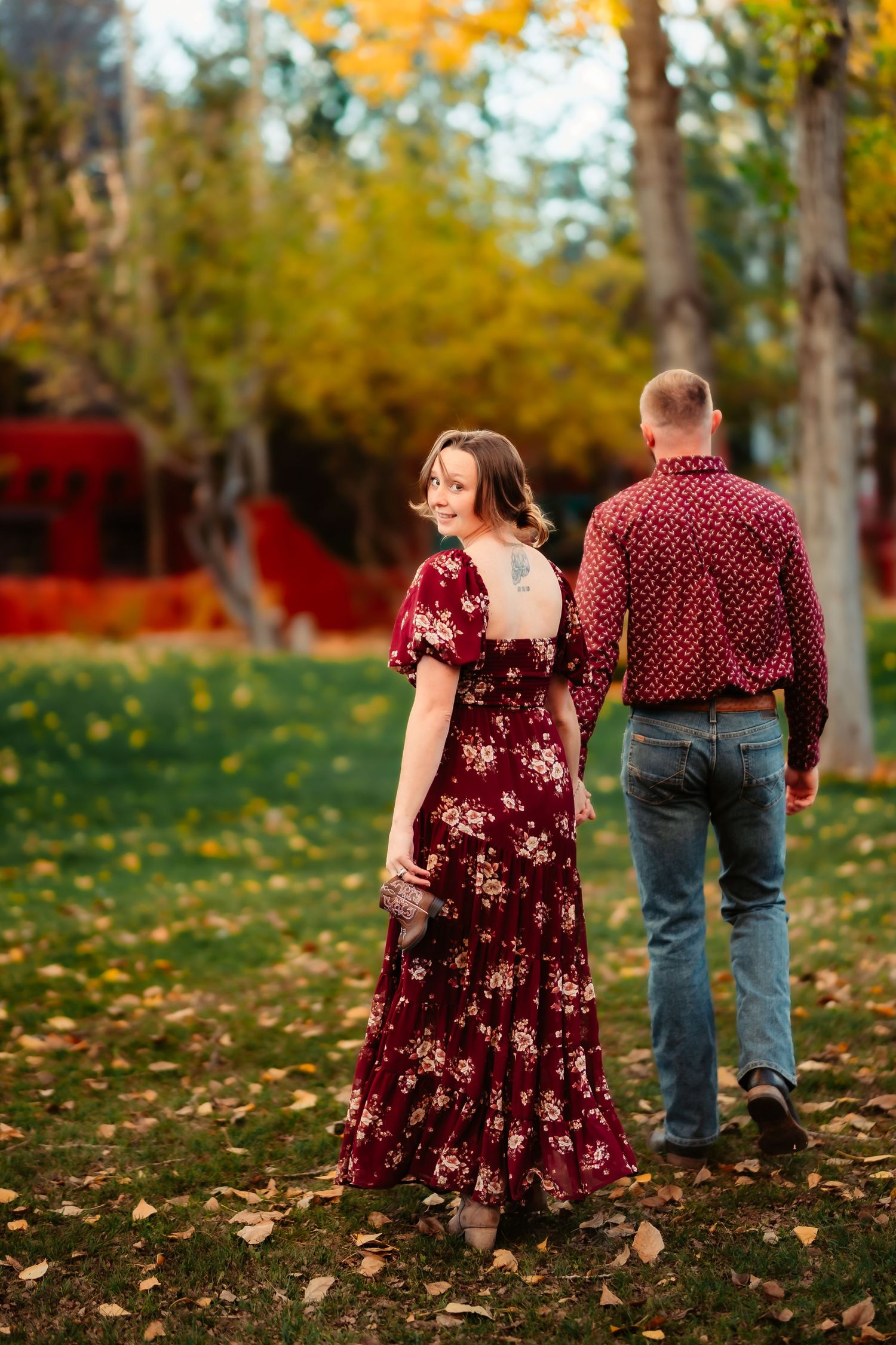 A couple walks through fallen autumn leaves in a park with burgundy and red clothing matching the fall foliage.