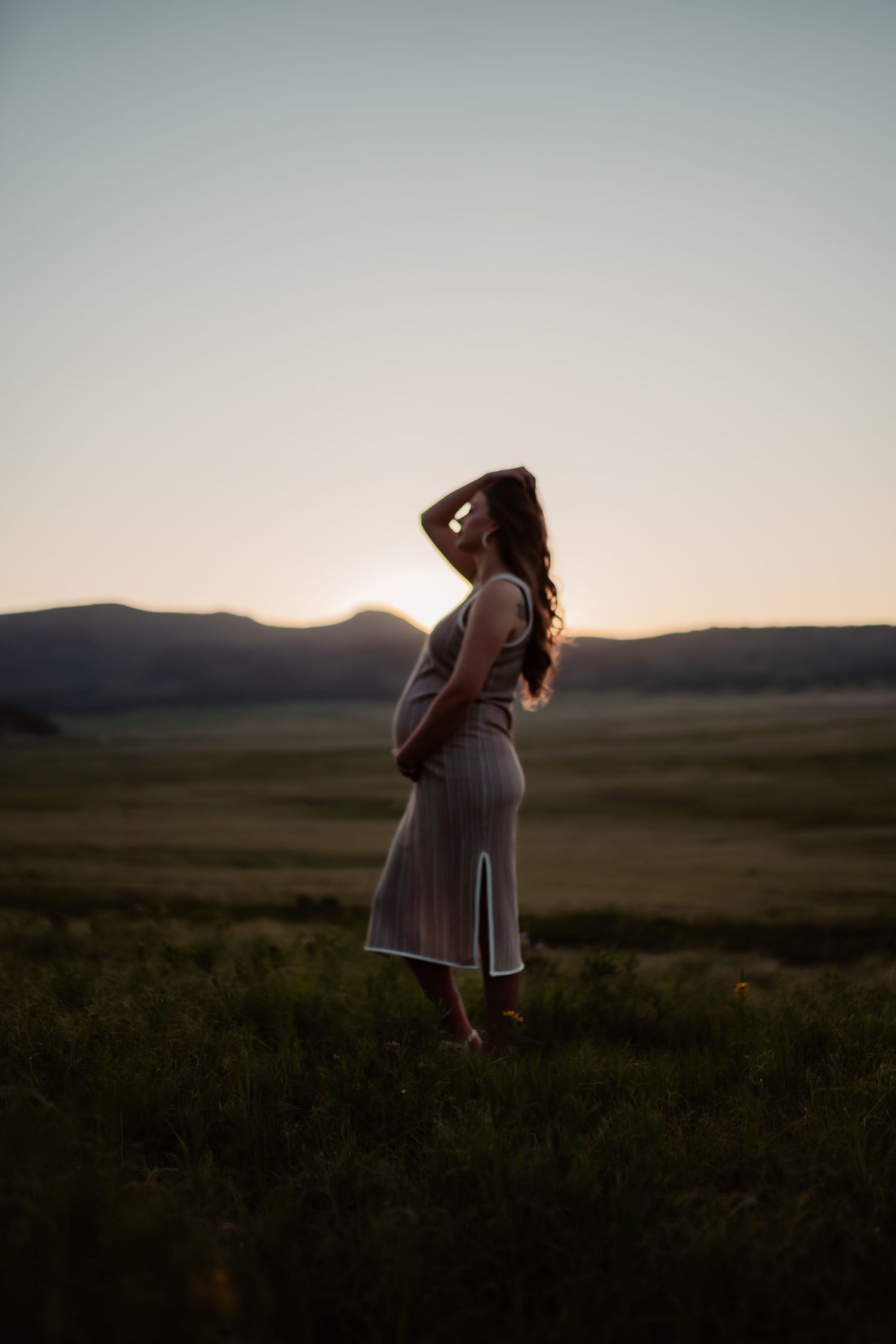 Silhouettes of a couple stand together against rolling hills and a dusky sky during golden hour.
