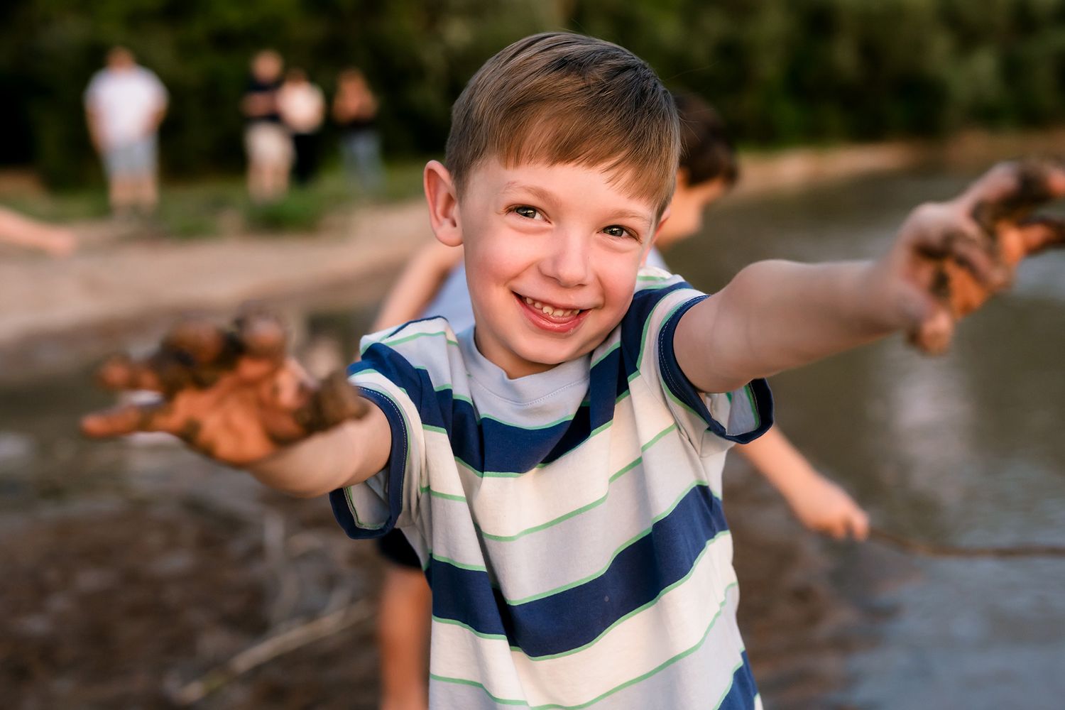 Smiling boy with muddy hands outstretched toward camera near a river, wearing a striped shirt outdoors.