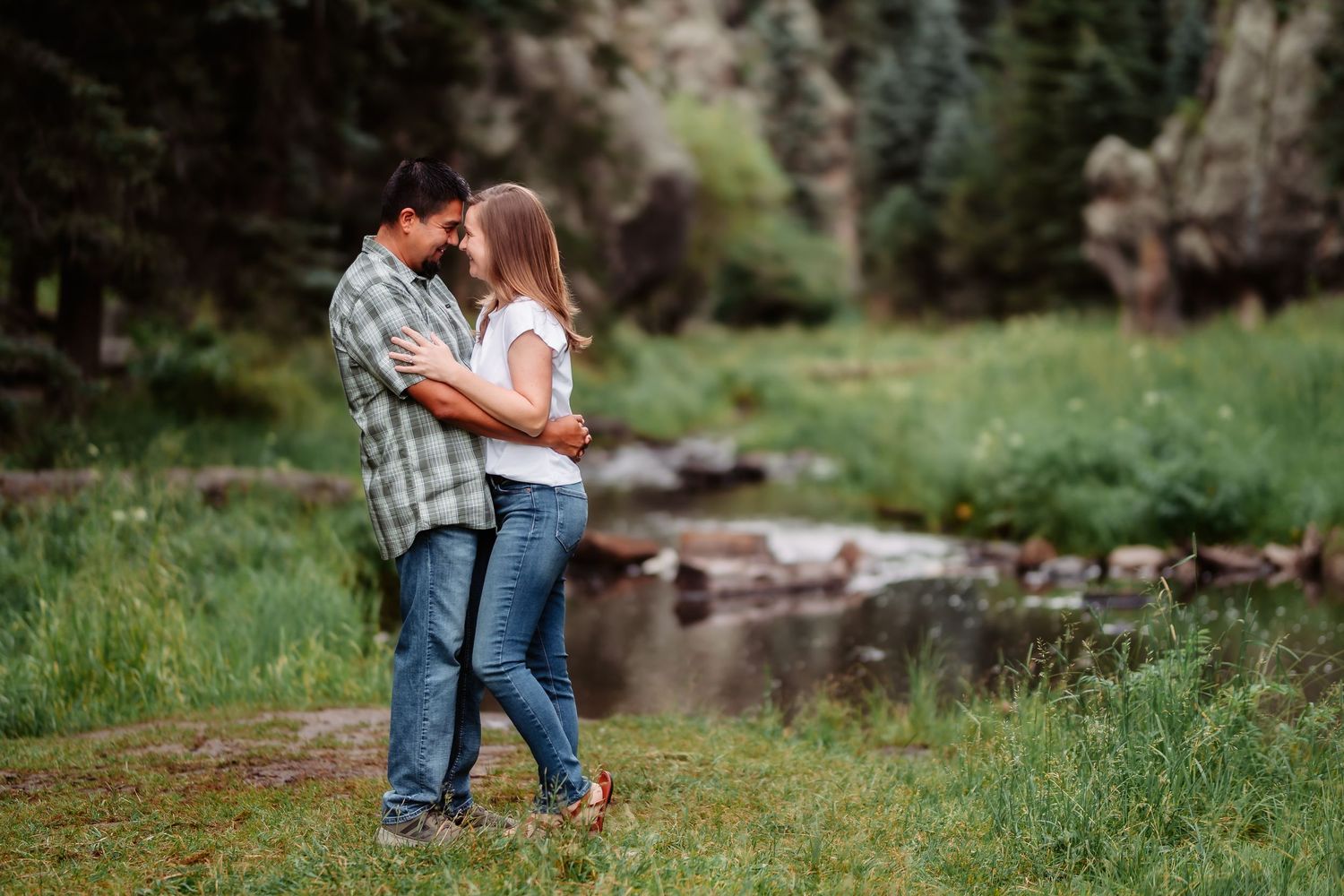 A couple embraces while standing in a scenic outdoor setting with trees and a stream in the background.