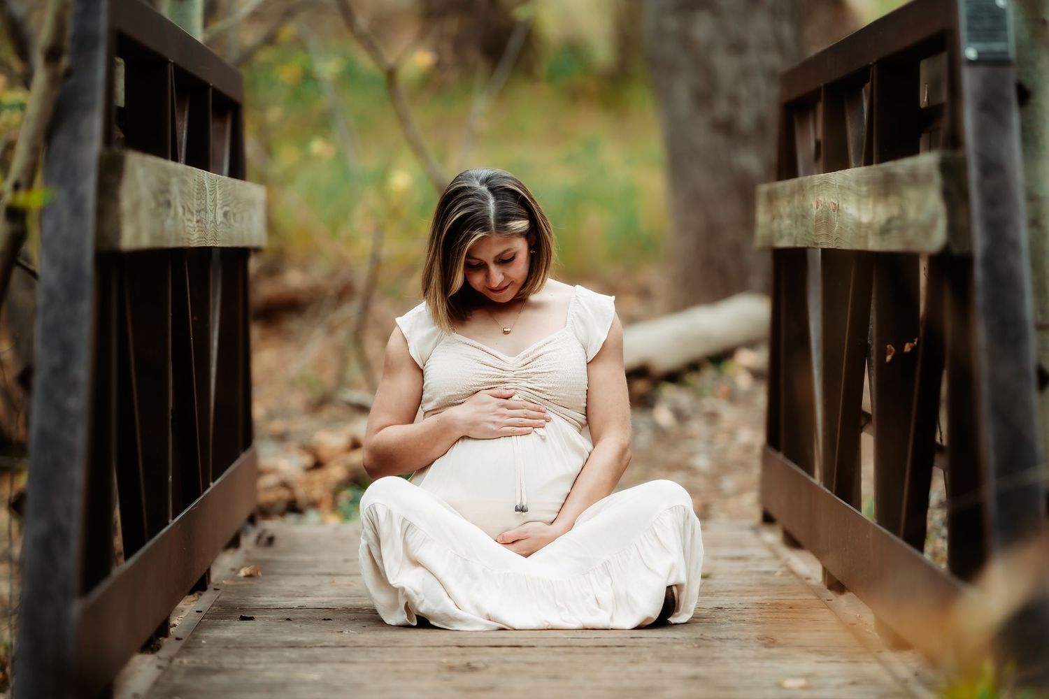 A person in a white dress sits on a rustic wooden bridge in a natural outdoor setting.