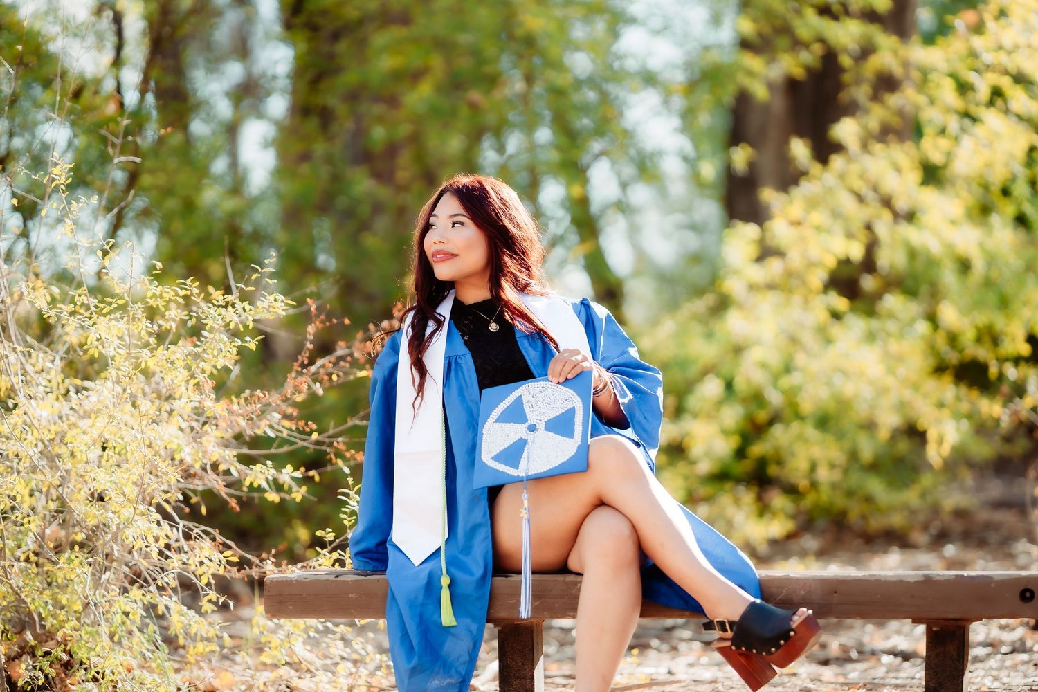 A person in a blue dress and white blazer poses outdoors among sunlit trees and foliage for a portrait session.