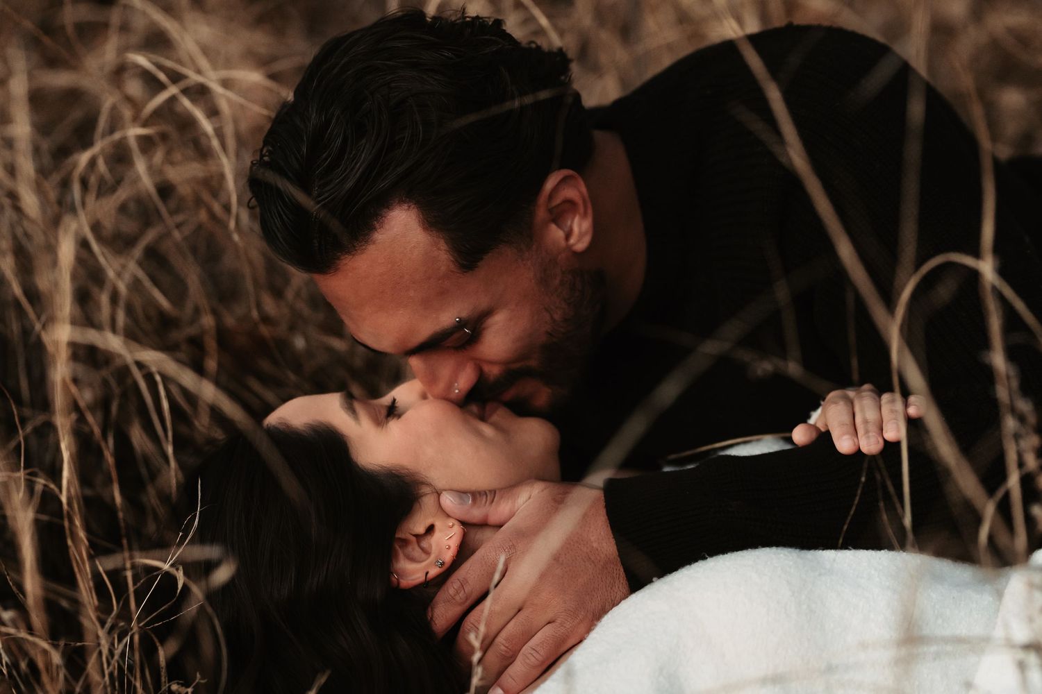Two people share a tender embrace while laughing together in a natural outdoor setting with dried plants.