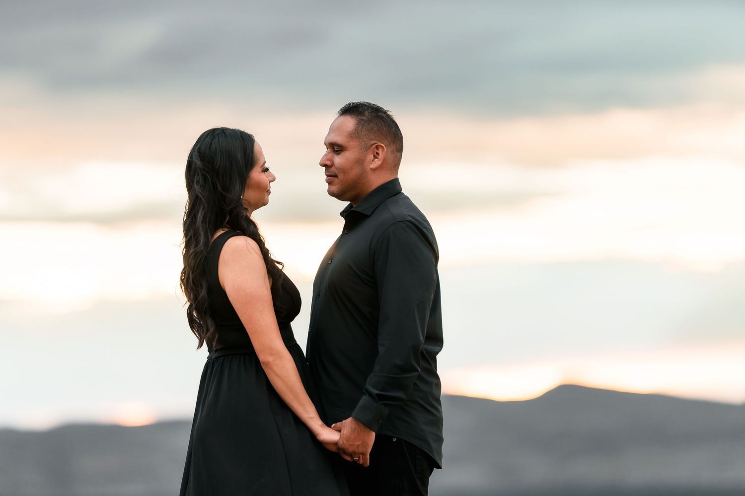 Couple in black holding hands facing each other against a pastel sunset desert sky.