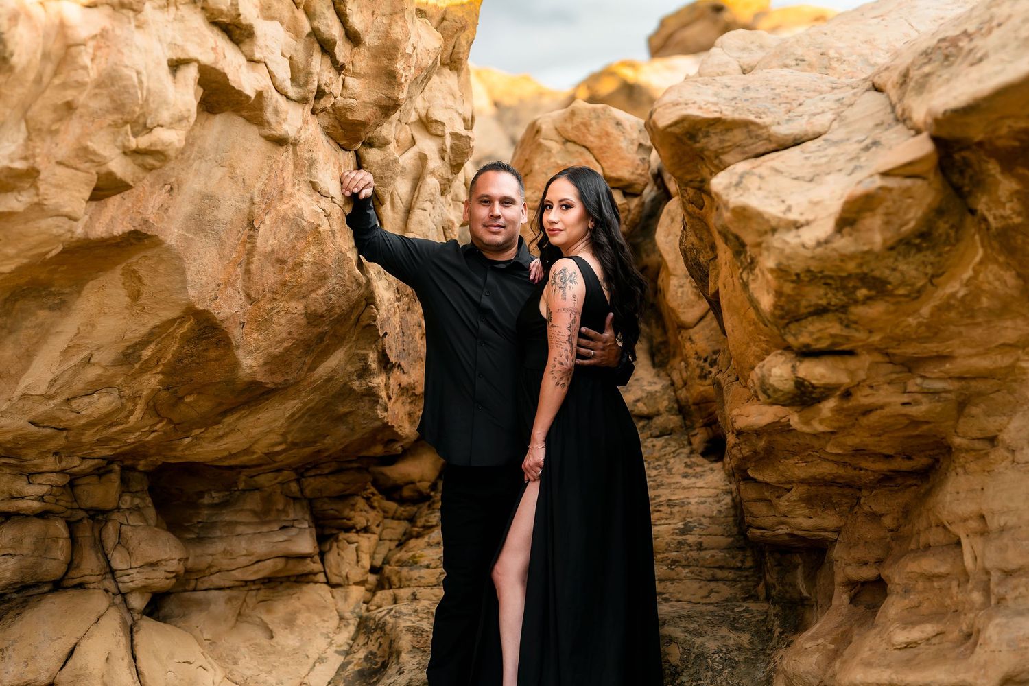 Couple in matching black outfits posing among golden sandstone rock formations in a desert landscape.