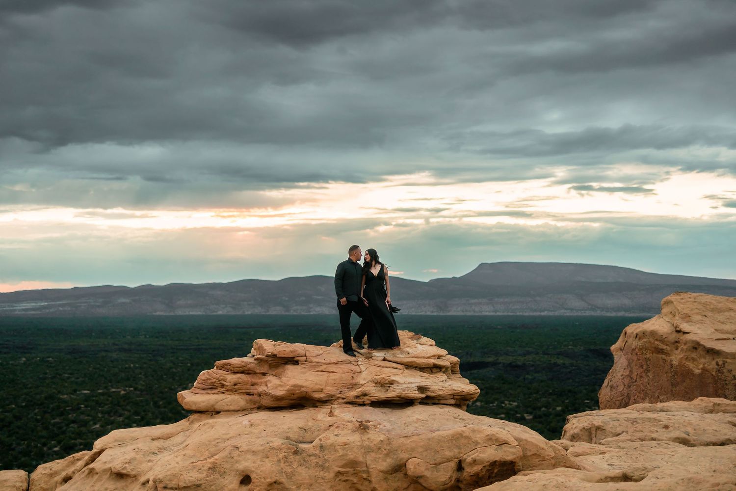 Couple in black outfits embracing on rocky desert cliff with vast green valley below.