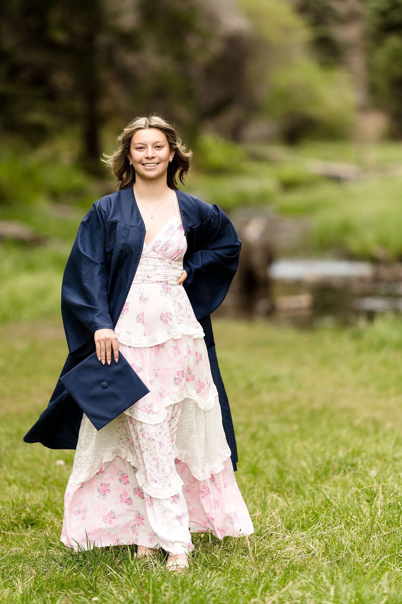 Smiling graduate in navy gown and floral dress holds cap while standing in green park.