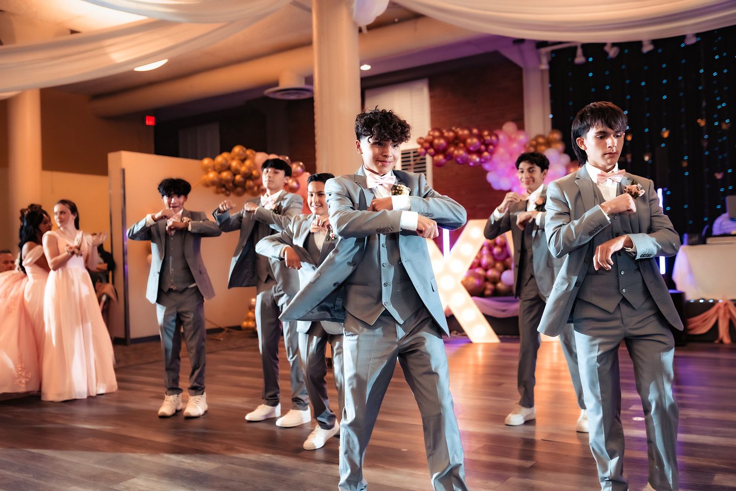 Wedding party performs a choreographed dance routine in gray suits and pink dresses at a reception venue.