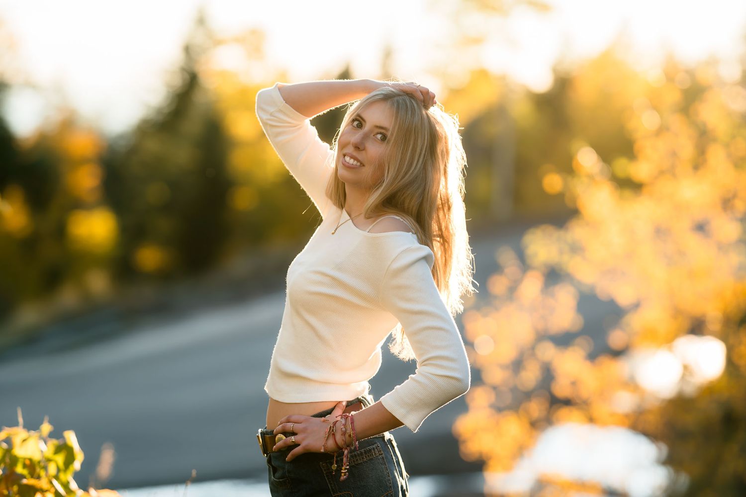 Young blonde woman in white crop top poses on a sunlit road surrounded by golden autumn foliage.