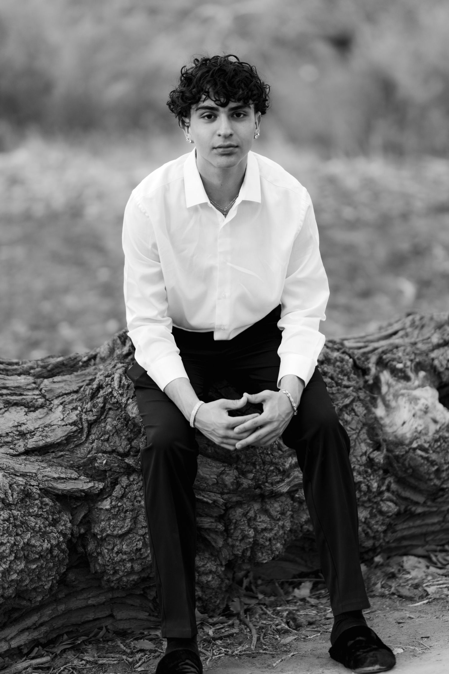 Black and white portrait of a person in white shirt sitting on a log with blurred natural background.
