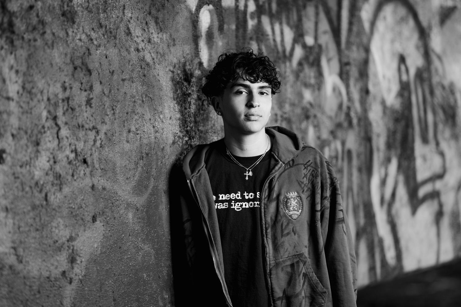 Moody black and white portrait against graffiti wall wearing dark jacket.