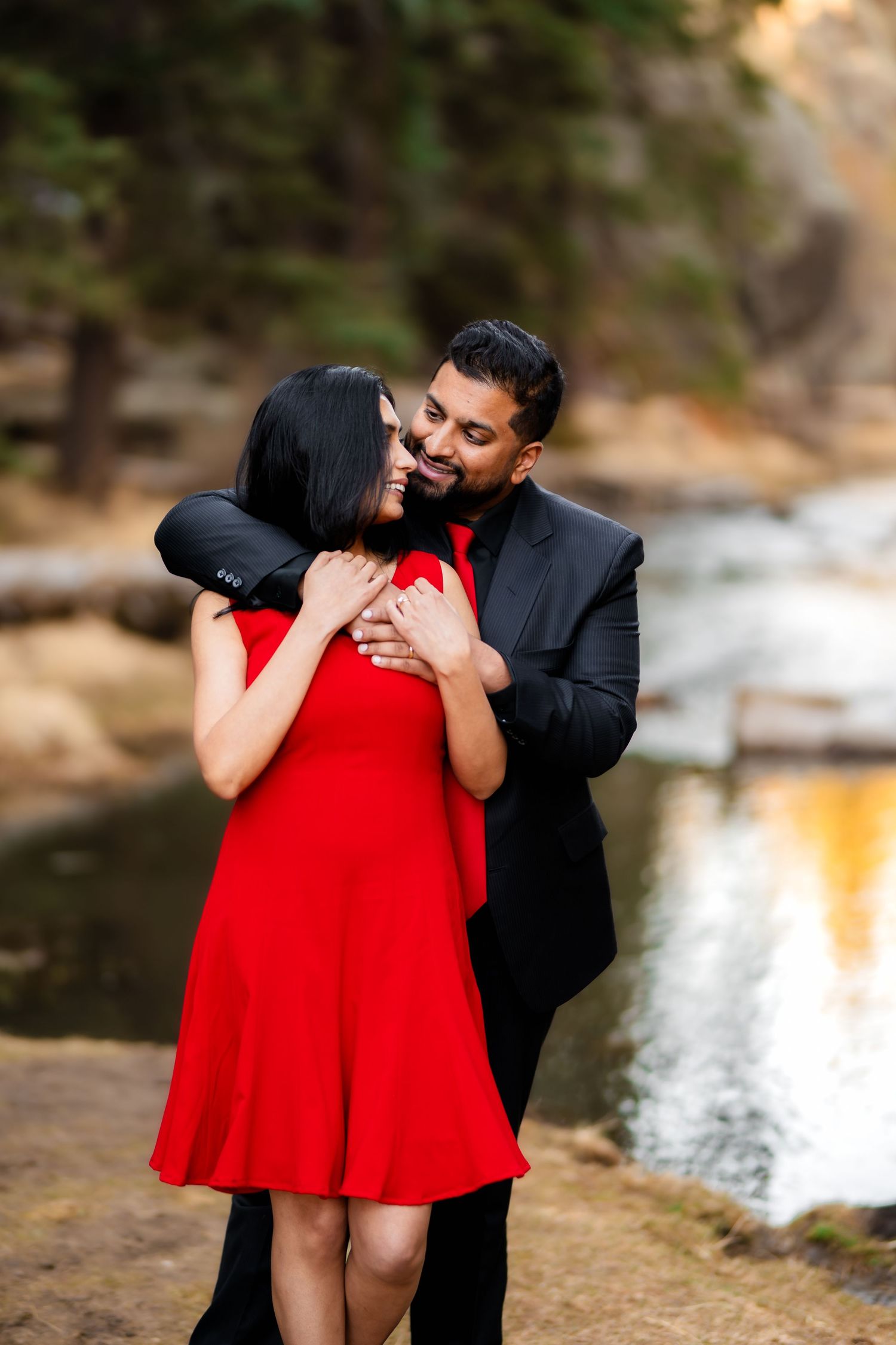 A romantic couple embraces near a waterfall, with the person in red dress and black suit sharing an intimate moment.