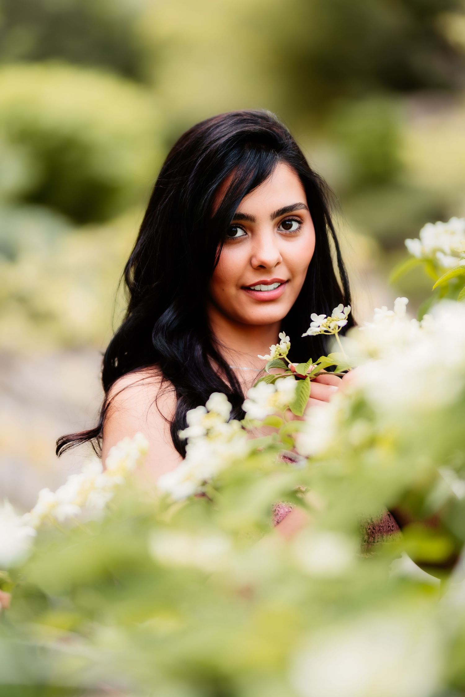 Portrait photograph of someone with long dark hair smiling among white flowers and greenery in a garden setting.