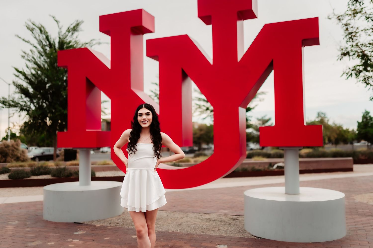 Large red UNM letters serve as iconic campus landmark at the University of New Mexico with student in white dress.
