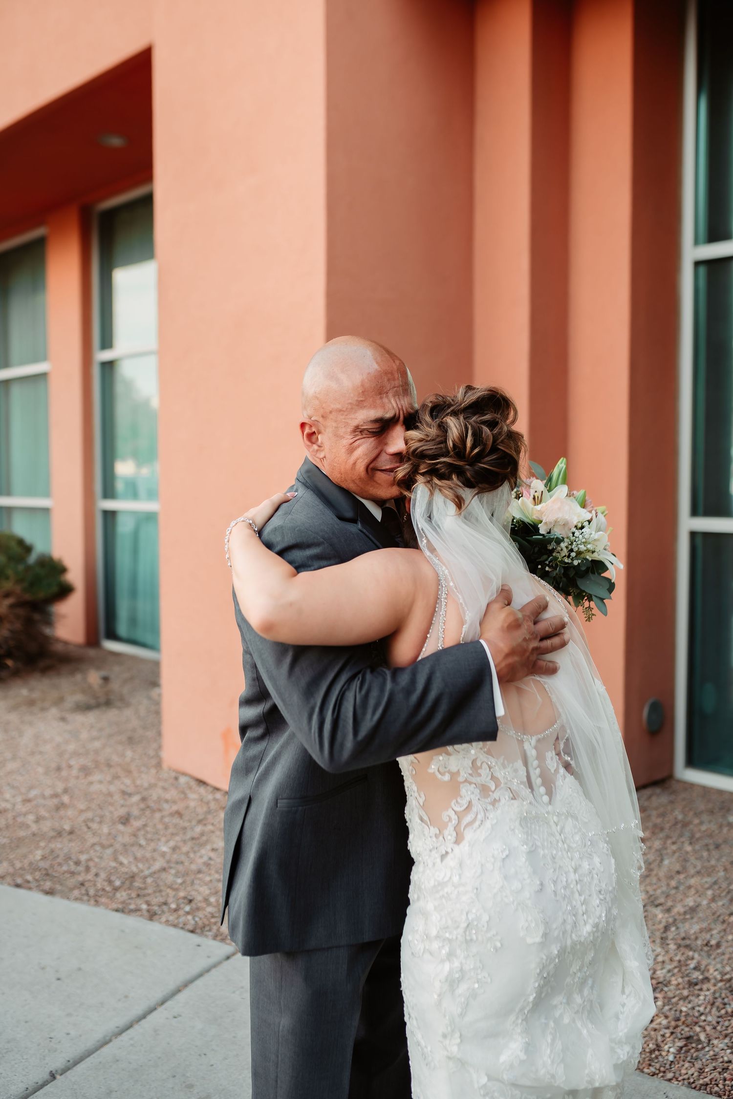Emotional first look moment captured with couple embracing outside a coral-colored building.